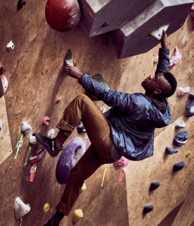 A person wearing a blue jacket and brown pants climbs an indoor bouldering wall, gripping colorful holds and reaching toward a large gray feature. The wall is covered with various climbing holds and route markers.