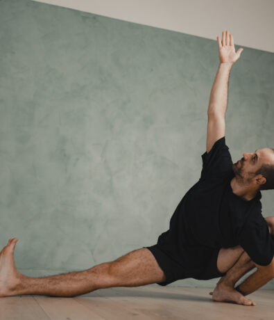 A person in a black outfit practices yoga indoors in Fremont, performing a deep side lunge with one arm extended upward and the other supporting the pose on the floor, set against upper walls painted green and a wooden floor.