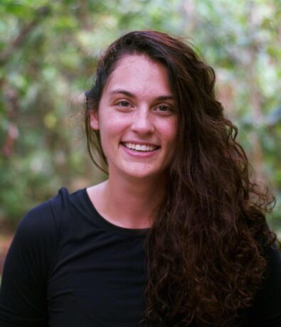 A young woman with long, curly brown hair and a black shirt smiles at the camera outdoors in Fremont, with green foliage and the Upper Walls blurred in the background.