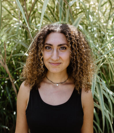 A young woman with curly brown hair and a nose ring stands in front of tall green grass in Fremont, wearing a black sleeveless top and layered necklaces, smiling softly at the camera.