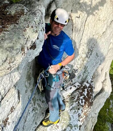 A climber in a helmet, blue shirt, and climbing gear—channeling Brooklyn Climbing Yoga and Fitness vibes—smiles while ascending a rocky cliff, with lush vegetation below.