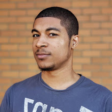 A young man with short hair and a trimmed beard wears a gray T-shirt and stands confidently in front of the brick Upper Walls in Fremont, looking directly at the camera.