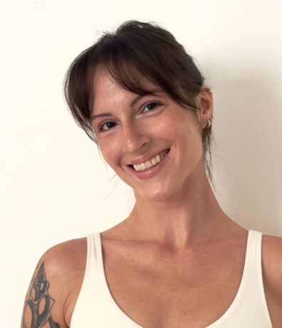 A woman with brown hair and bangs, wearing a white tank top, smiles at the camera. She has a tattoo on her right shoulder and stands in front of a plain white background, ready for a Brooklyn Climbing Yoga and Fitness session.