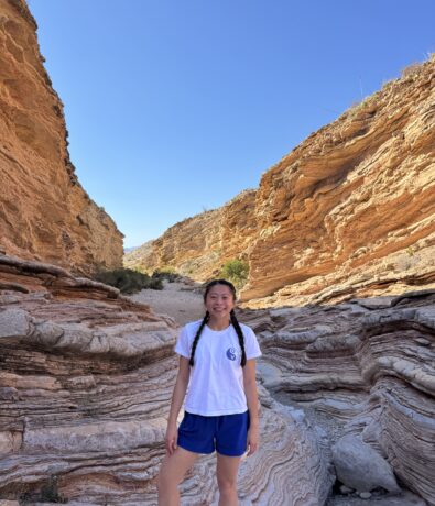 A young woman with braided hair, wearing a white T-shirt and blue shorts, stands smiling in a rocky canyon with layered orange rock formations under a clear blue sky.