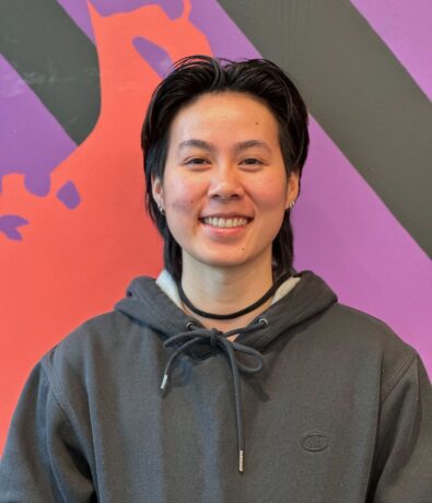 A person with short dark hair, wearing a gray hoodie, smiles in front of a colorful wall with purple, orange, and black geometric shapes at Brooklyn Climbing Yoga and Fitness.