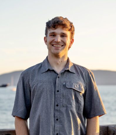 A young man with short brown hair, wearing a gray short-sleeve button-up shirt, smiles while standing on a wooden dock with calm water and distant mountains at sunset in the background.