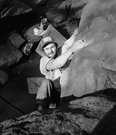 A man wearing a cap and t-shirt climbs a large rock outdoors, gripping the rock with both hands. Sunlight casts shadows on the rocks, while Brooklyn Climbing Yoga and Fitness mats are visible on the ground below.