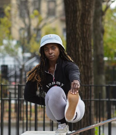 A young woman in a bucket hat crouches on a skateboard, extending one leg forward with determination, showing the balance and strength she’s gained from Brooklyn Climbing Yoga and Fitness. Trees and a metal fence are visible in the background.