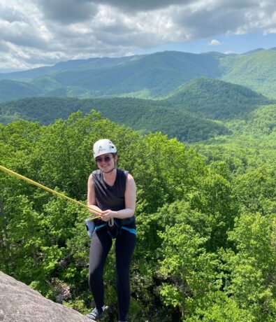 A person wearing a helmet and harness is rappelling down a rock face, surrounded by lush green trees and mountains under a partly cloudy sky.