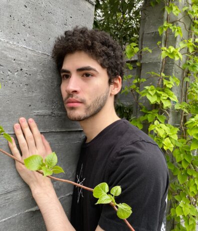 A young person with curly hair and a short beard, wearing a black t-shirt, stands next to a concrete wall partially covered with green ivy outside Brooklyn Climbing Yoga and Fitness, gazing thoughtfully into the distance.