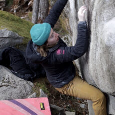 A person wearing a teal beanie, black jacket, and brown pants is bouldering on a rock face outdoors, reaching upward with one hand. A crash pad lies on the ground below.