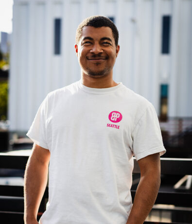 A man stands outdoors, smiling at the camera in a white T-shirt with a pink logo and SEATTLE printed on it—perfect for showing off your summer membership style against a backdrop of a modern white building and lush greenery.