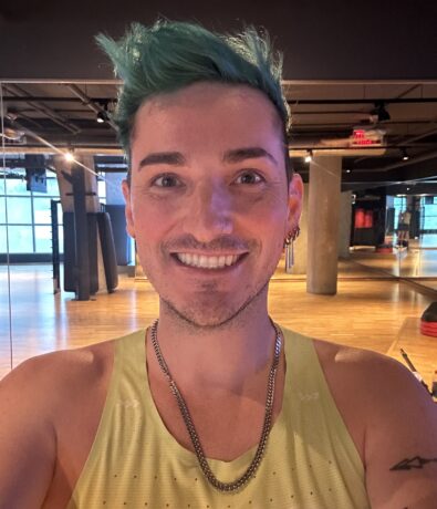 A person with short green hair and a trimmed beard smiles at the camera in a spacious, modern gym with climbing walls, wooden floors, large windows, and mirrored walls. They are wearing a light yellow tank top and a silver chain necklace.