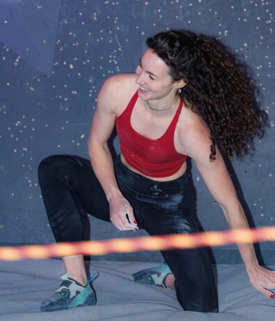 A woman with curly hair, wearing a red sports bra, black leggings, and climbing shoes, kneels and smiles on an indoor climbing mat in Tempe, with a climbing wall and holds visible in the background—perfect for personal coaching sessions.