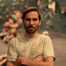 A man with short brown hair and a beard stands with his arms crossed and smiles slightly. He is wearing a light-colored t-shirt at an indoor climbing gym with blurred people and climbing walls in the background.