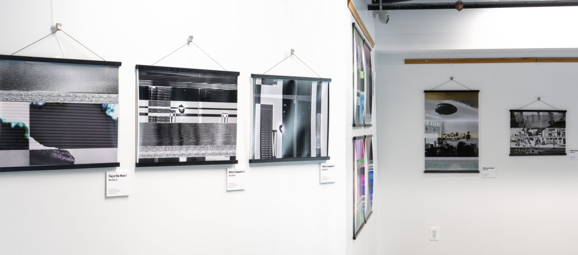 Four abstract black and white photographs hang on white gallery walls at a DC arts space, each with a label beneath. A hallway leads to more framed artwork, with pipes and an exposed ceiling visible above. Part of the Community Arts Program.