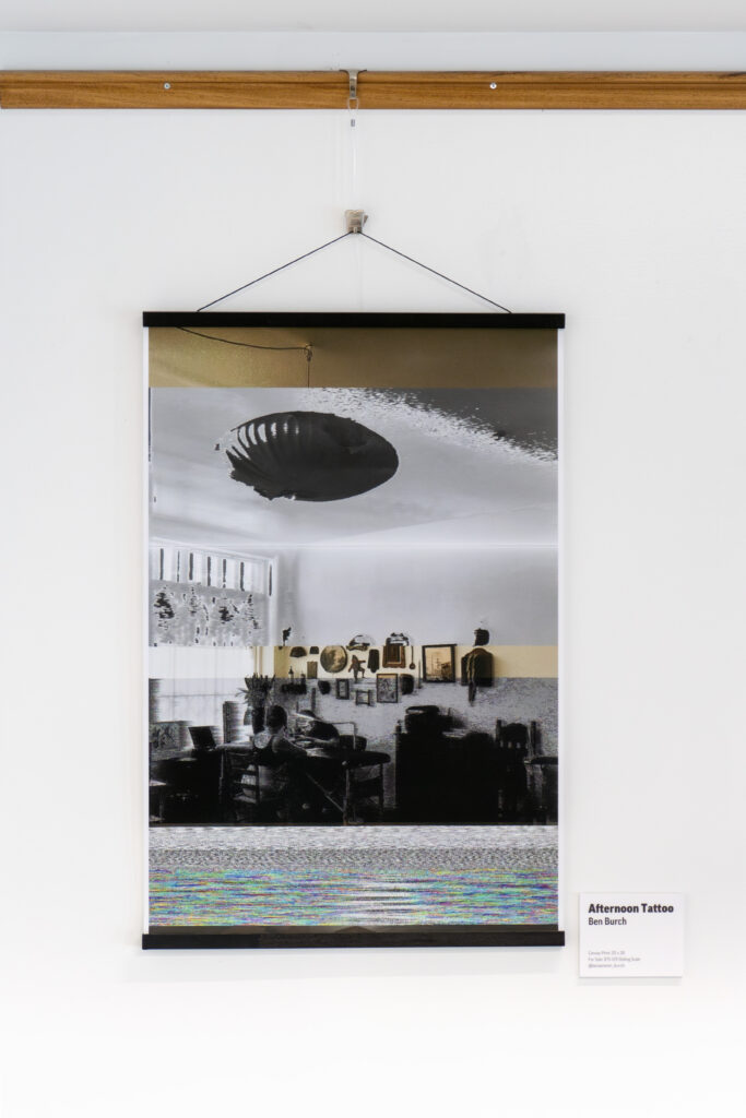 A framed artwork titled Afternoon Tattoo hangs on a white wall, showing a stylized, partially distorted black-and-white photo of a room with furniture and decorations. Featured as part of the DC Arts Program, it includes a small label on the lower right side.