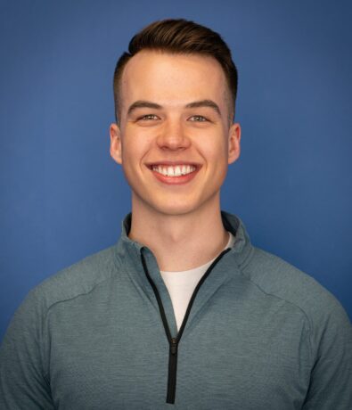 A young man with short brown hair smiles, wearing a light blue zip-up pullover over a white shirt, standing in front of a plain blue background, ready for a Personal Training session in the Twin Cities.