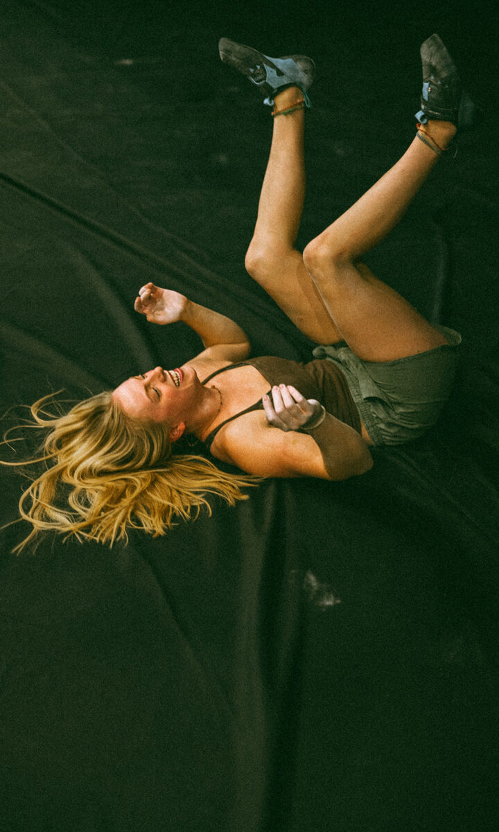 A woman with long blonde hair lies on her back, smiling with arms bent and legs raised, in a st paul rock climbing gym. She wears a tank top, shorts, and climbing shoes on the dark gym surface.