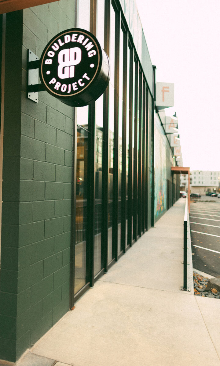 A round, illuminated sign reading Bouldering Project is mounted on a green brick building with large windows, marking this spot as a St Paul rock climbing gym next to the sidewalk and a mostly empty parking lot.