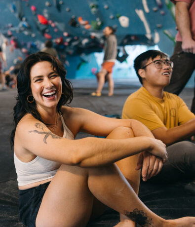 A smiling woman sits on the floor of Climbing Tempe’s indoor gym, arms resting on her knees. Nearby, a man in a yellow shirt relaxes as colorful climbing holds fill the background.