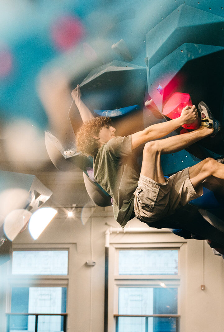A person with curly hair climbs an indoor bouldering wall at a climbing gym in the University District, Seattle, gripping colorful holds. The image features reflections and blurred foreground, with windows and lights in the background.