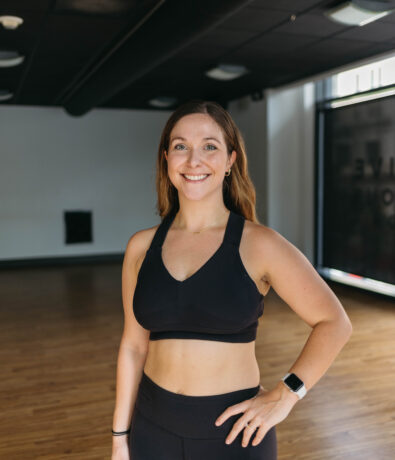 A woman in athletic wear stands confidently in a gym with wooden floors and large windows, smiling with one hand on her hip—ready for her next climbing session.
