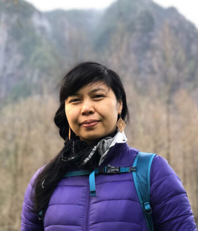 A woman in a purple jacket and patterned earrings stands outdoors near the Upper Walls in Fremont, with a blurred backdrop of trees and rocky mountains. She has a slight smile and carries a backpack strap over her shoulder.