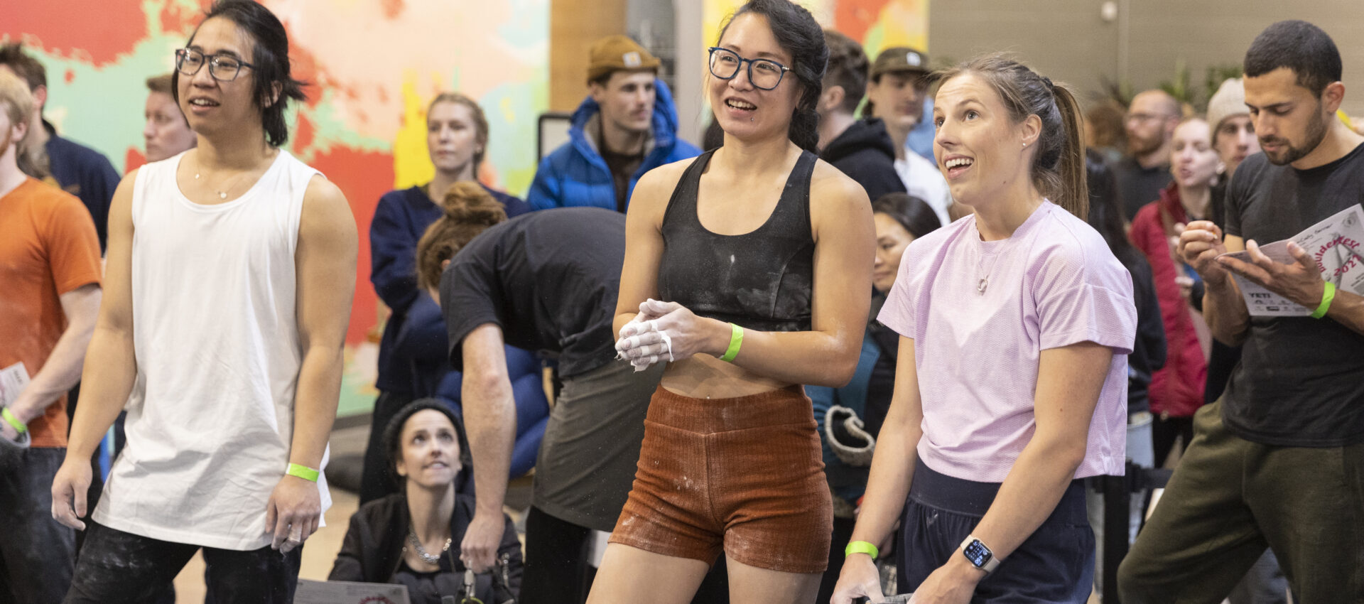 Three people stand in the foreground of a rock climbing gym in Minneapolis, smiling and looking up, with a crowd of onlookers and colorful walls behind them. All wear athletic clothing and have chalk on their hands, reflecting the spirit of local community clubs.