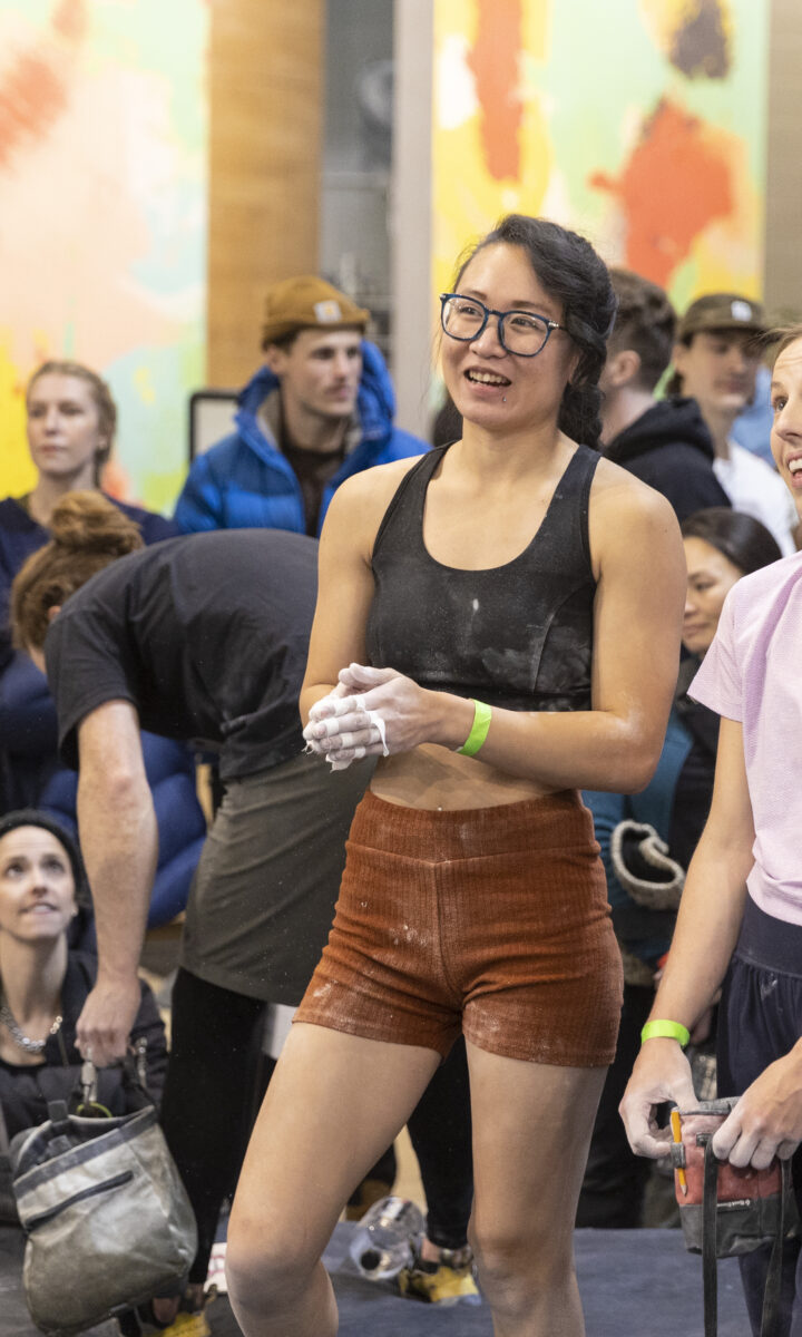 Three people stand in the foreground of a rock climbing gym in Minneapolis, smiling and looking up, with a crowd of onlookers and colorful walls behind them. All wear athletic clothing and have chalk on their hands, reflecting the spirit of local community clubs.