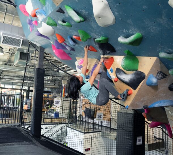 A woman in athletic clothing is climbing an indoor bouldering wall with colorful holds, using her hands and feet to grip an overhanging section. The gym features mats, nets, and industrial elements.