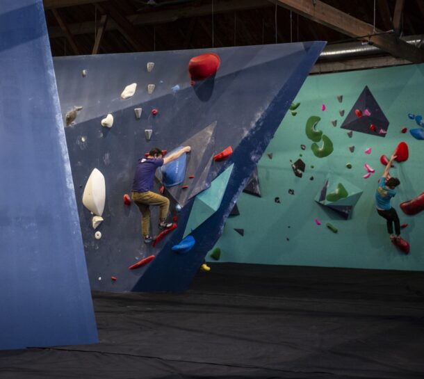Two people are indoor rock climbing on blue and green artificial climbing walls at Upper Walls in Fremont, with colorful holds. The floor is padded for safety, and exposed wooden beams run above the climbing area.