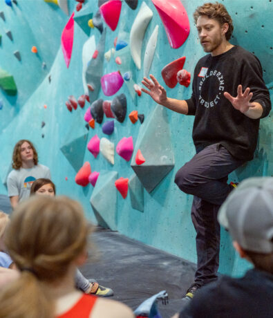 A man leans against an indoor climbing wall, speaking to a group of people seated on the floor, who are listening attentively. The wall is covered with colorful climbing holds.