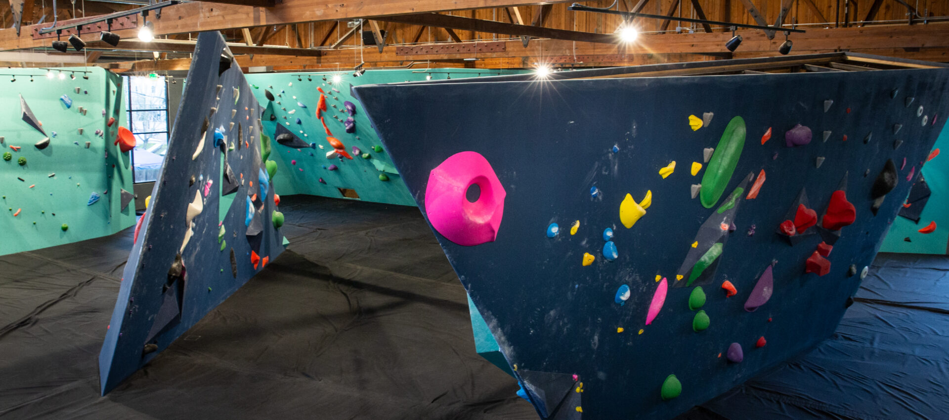 Indoor bouldering gym in Fremont featuring angular climbing walls from Upper Walls, covered in colorful holds on padded flooring, all set within a building with exposed wooden beams and natural light from windows.
