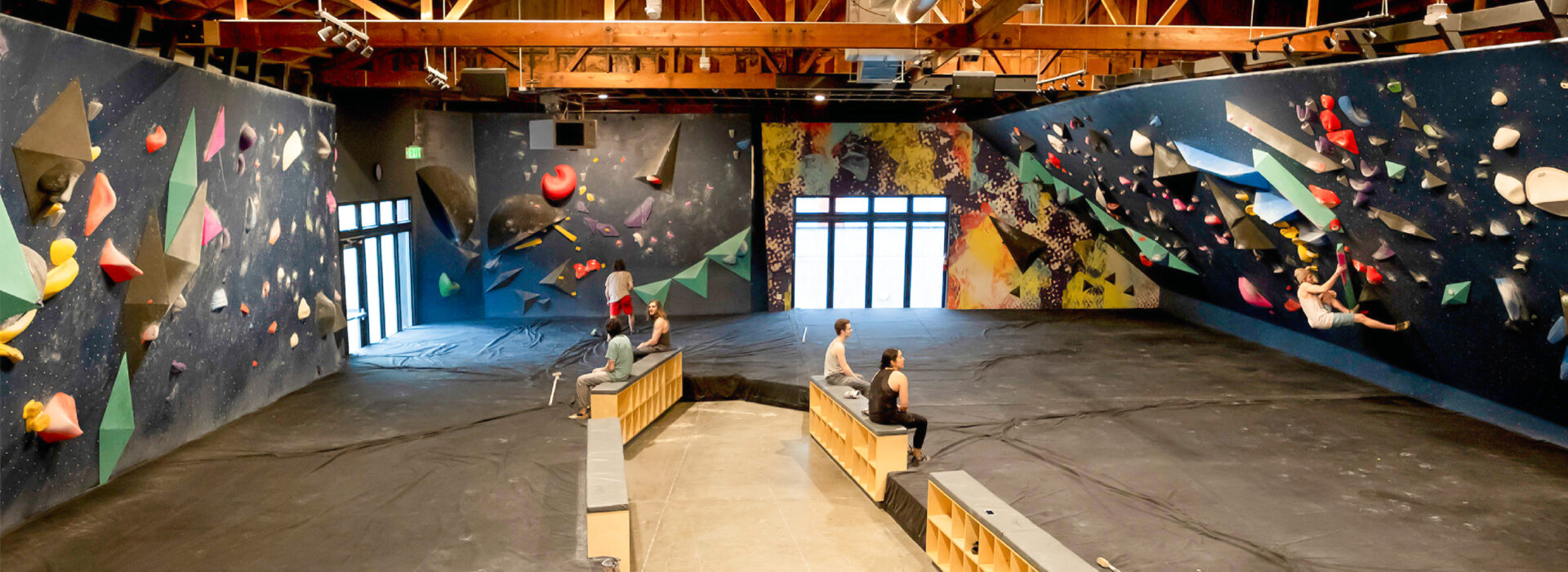 Indoor bouldering gym in Fremont with colorful climbing holds on angled Upper Walls, wooden ceiling beams, and a few people sitting or climbing. Natural light enters through large windows at the far end.