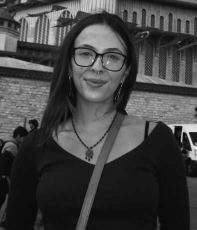 A woman with long hair, glasses, and a nose ring stands outdoors in front of a large, ornate building. She is wearing a necklace and a shoulder bag, smiling slightly at the camera. The image is in black and white.