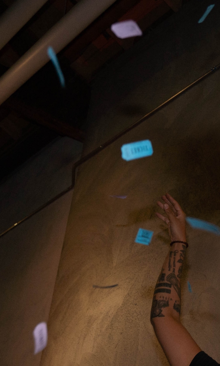 A tattooed arm reaches upward as blue raffle tickets or pieces of paper fall in the air against a dimly lit wall and ceiling.