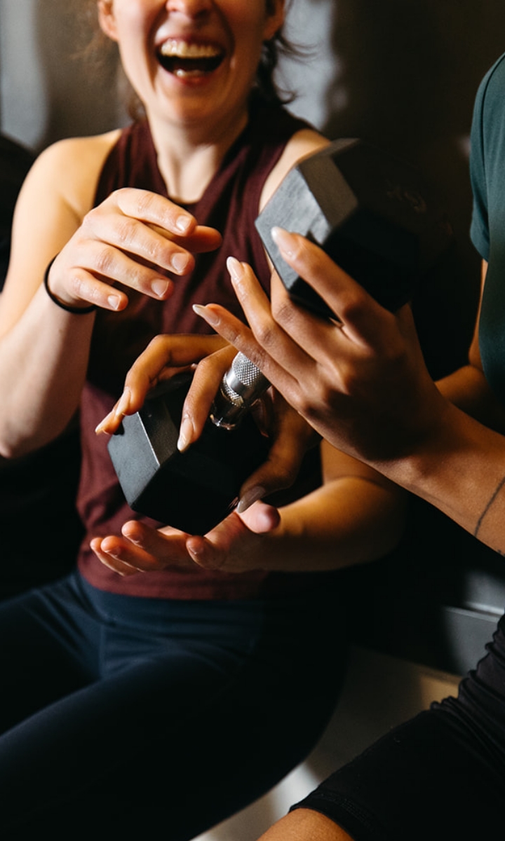 Two people smiling and laughing while holding a dumbbell together, focusing on their hands and the weight—one with a bandaged hand. The friendly, supportive atmosphere captures the spirit of Fitness Classes.