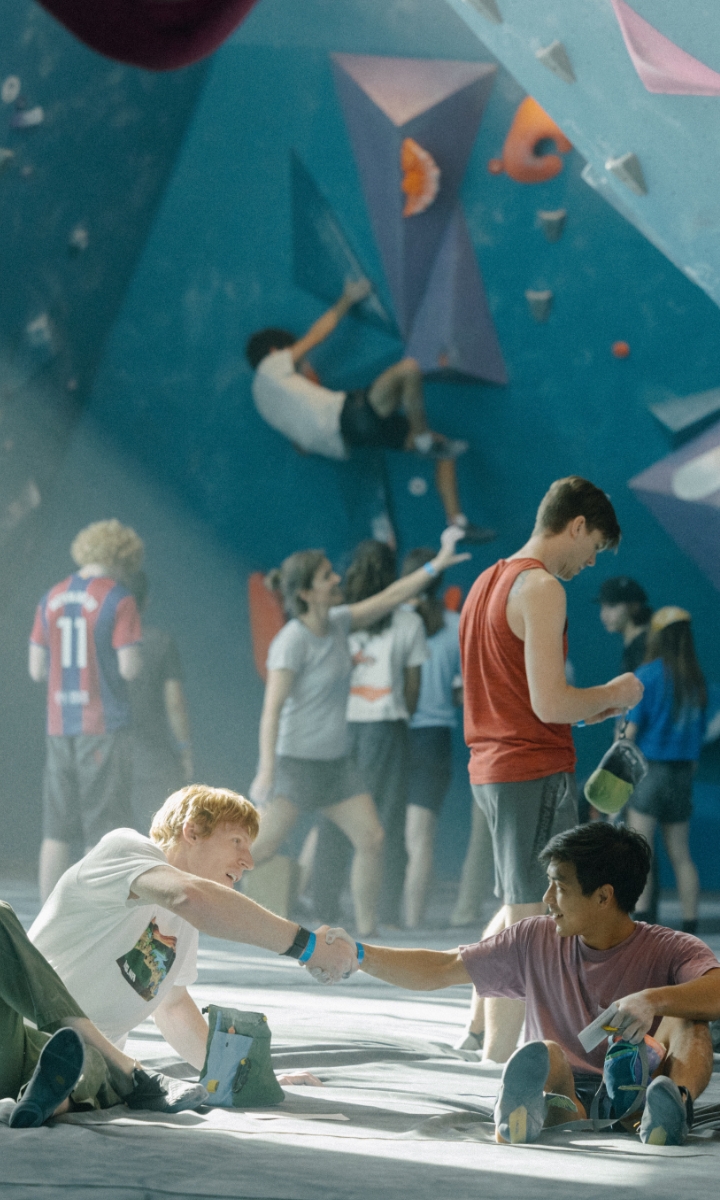 Two young men sitting on the floor of an indoor rock climbing gym shake hands and smile, while climbers scale colorful walls and others socialize in the background. Sunlight filters through the room, creating a lively atmosphere.