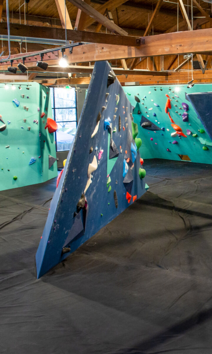 A modern indoor climbing gym in Fremont featuring blue and green angled climbing walls, colorful holds, Upper Walls for advanced climbers, and padded black flooring—all under a wooden ceiling with exposed beams and bright lighting.