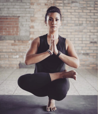 A woman practices yoga indoors, balancing on one leg with the other leg crossed over it. She holds her palms together at her chest, wearing black workout clothes, and is focused in a meditative pose against a brick wall background.