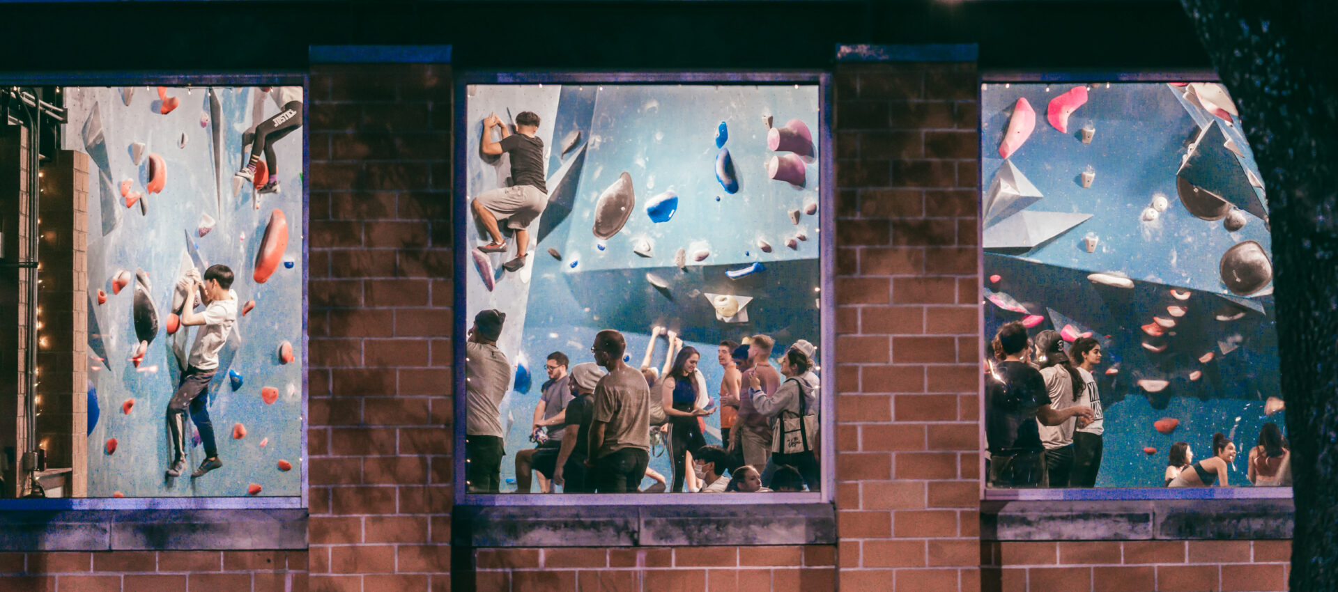 People are visible through large windows at an indoor rock climbing gym, some scaling climbing walls while others watch or wait below. The scene is lively, with colorful climbing holds and a crowd inside.