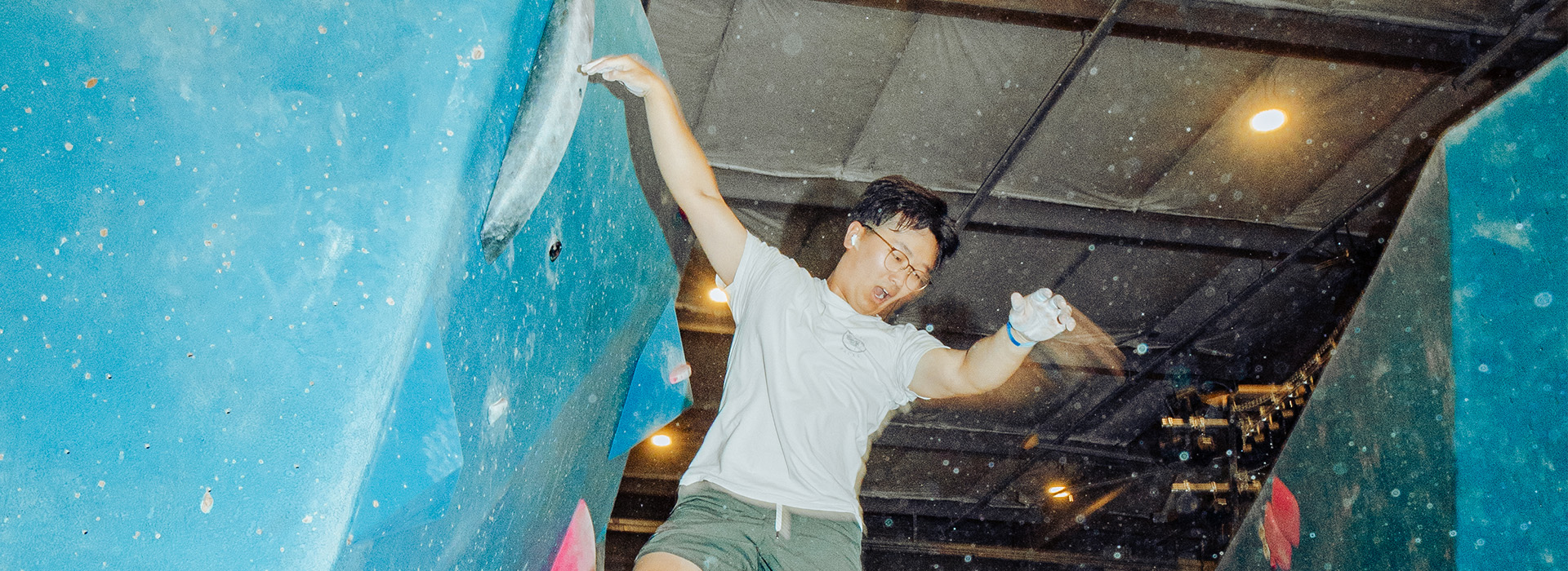 A person wearing glasses and a white shirt is climbing an indoor bouldering wall at Austin Bouldering Project, reaching for a hold with one hand and chalk on their hands—enjoying the Membership Benefits that support every determined climb.