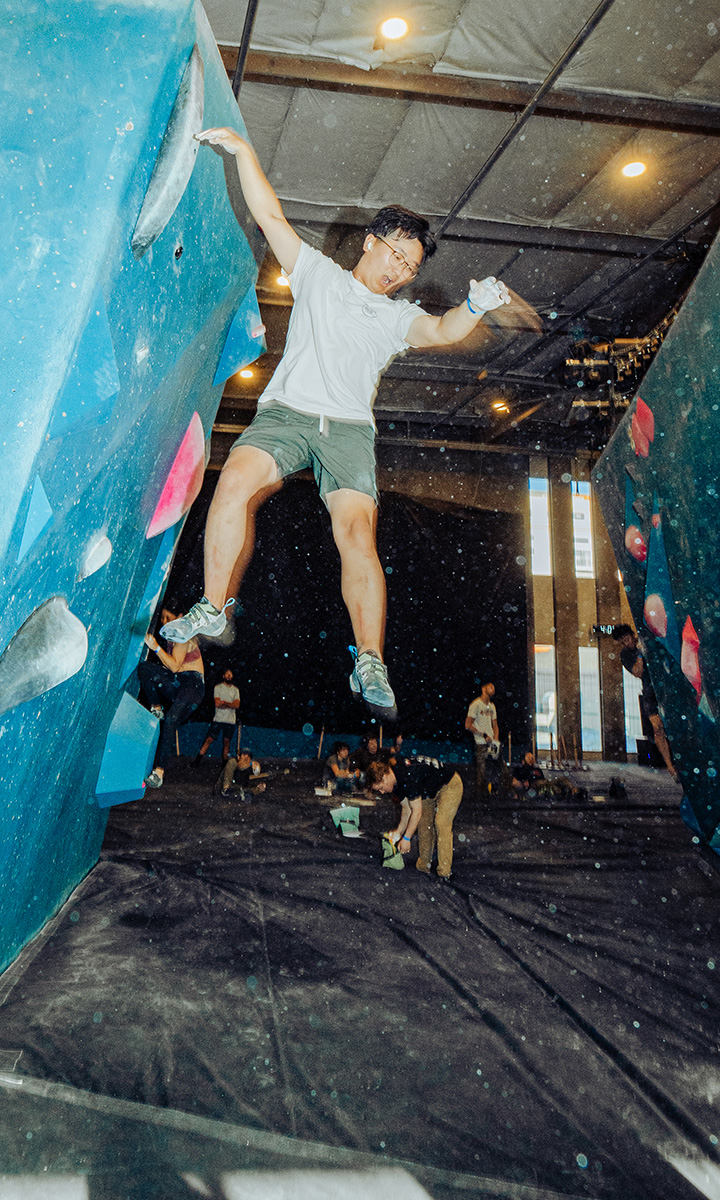 A person wearing a white shirt and green shorts is jumping off an indoor climbing wall at Seattle Bouldering Project, where membership benefits include access to blue panels, pink holds, and a vibrant community of climbers watching or climbing nearby.