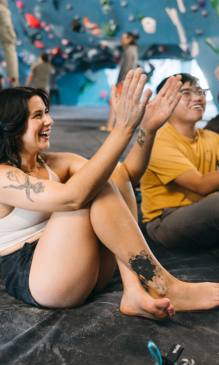 Two people sit on a padded gym floor, smiling and giving each other a high five. Colorful climbing holds are visible on the wall behind them. Enjoy moments like these with Membership Benefits at Seattle Bouldering Project. One person has tattoos and is barefoot.