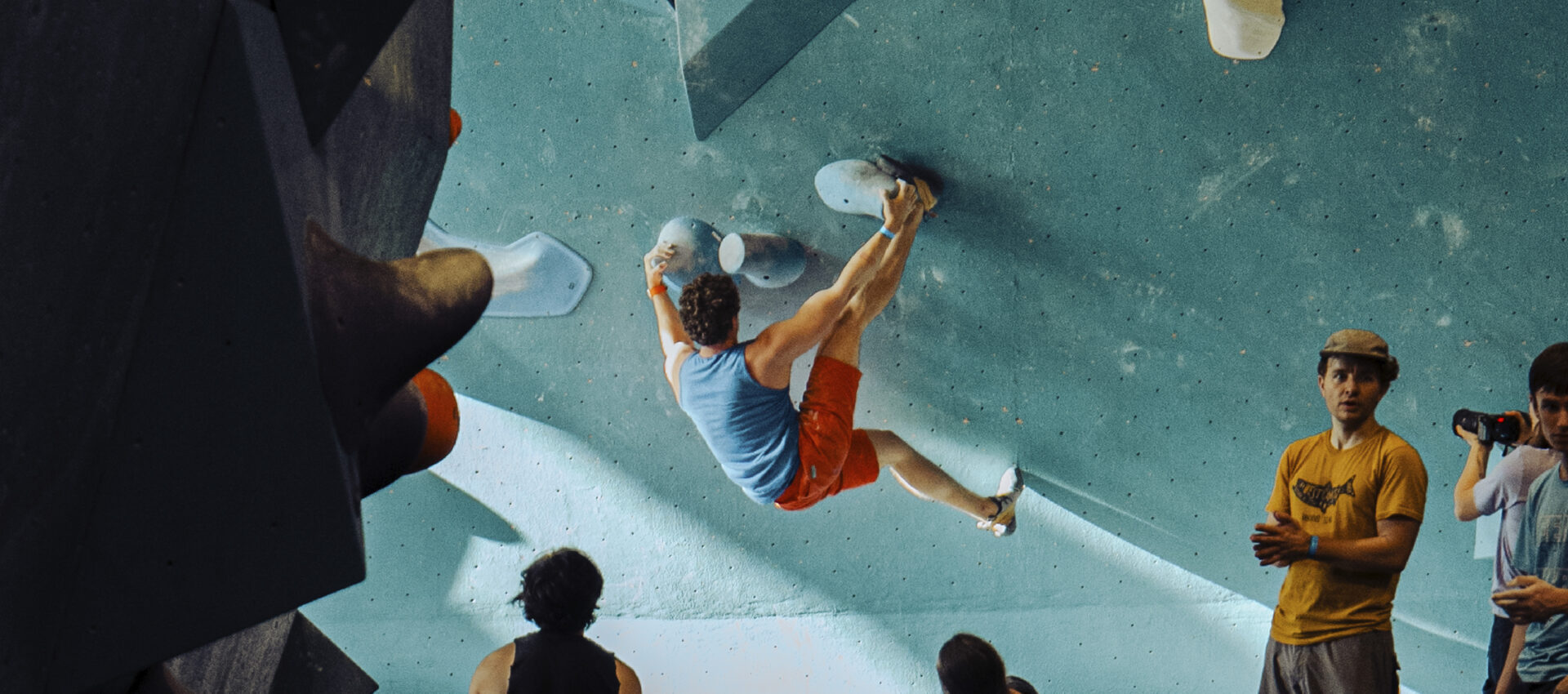 A man in a blue tank top and orange shorts climbs an indoor bouldering wall with colorful holds, as a group—some chatting about the new Membership Freeze option—watches from below, some standing and others sitting.
