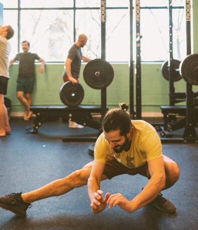 A man with a beard and tied-back hair stretches on the gym floor, while three others stand and talk near weightlifting racks in the background. Bright sunlight comes through the windows.