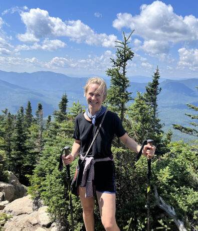 A person in hiking gear stands on a rocky mountain trail surrounded by green trees, holding trekking poles, with blue mountains and a bright, partly cloudy sky in the background.