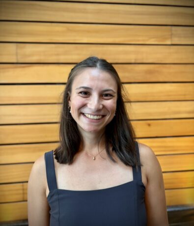 A woman with straight brown hair in a sleeveless navy dress smiles in front of a background of horizontal wooden panels.