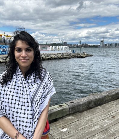 A woman with dark curly hair stands on a wooden dock by the water, wearing a black and white patterned scarf. Behind her are rocks, a sign that reads “ACAVE”, buildings, and a cloudy sky.
